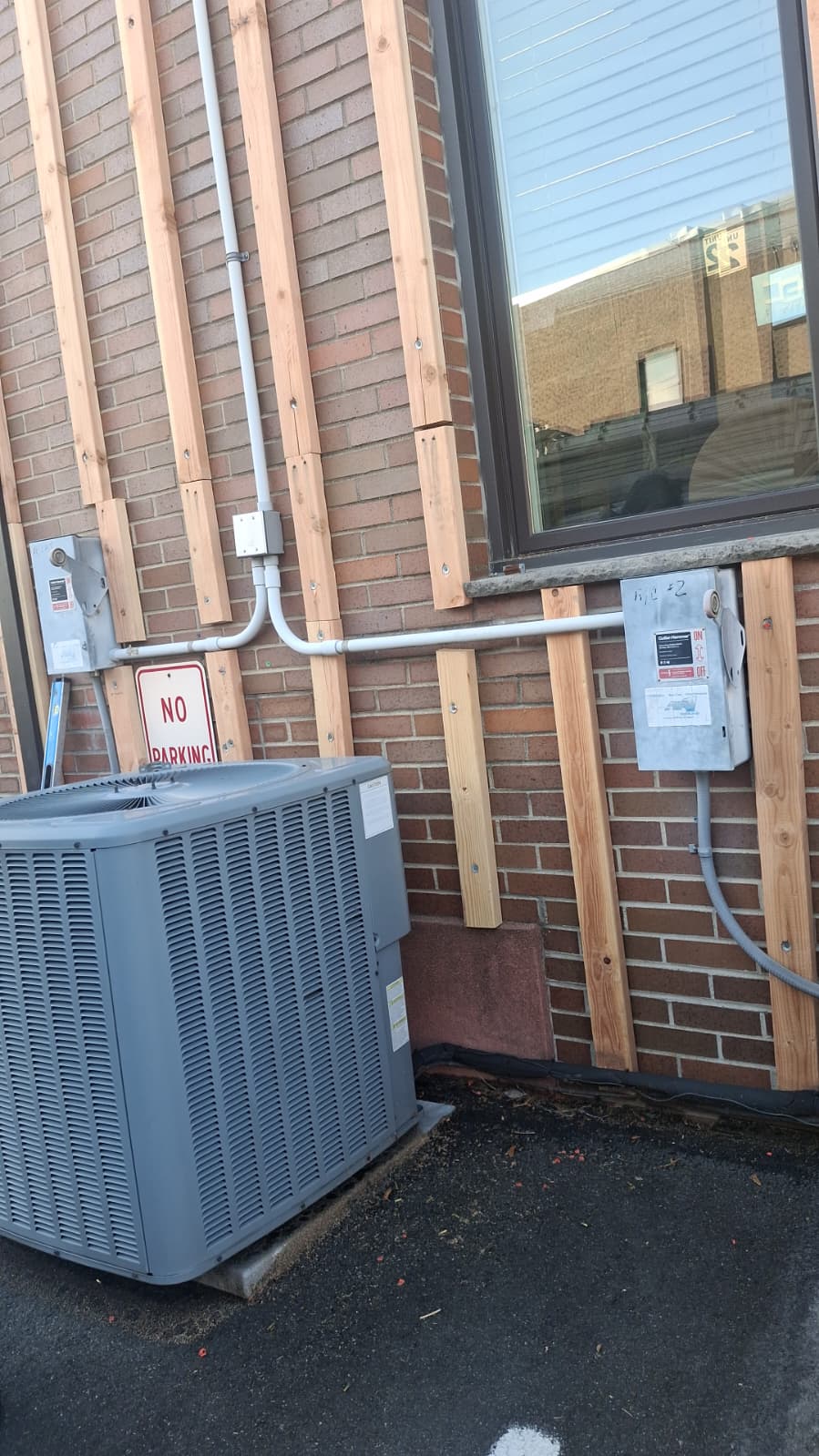 Close-up of vertical 2x4 furring laid out at 16 inches on center over brick, routed cleanly around AC condenser and electrical disconnect
